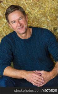 Portrait shot of an attractive, successful and happy middle aged man male wearing a blue sweater sitting on hay bales in a barn or stables