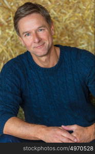 Portrait shot of an attractive, successful and happy middle aged man male wearing a blue sweater sitting on hay bales in a barn or stables