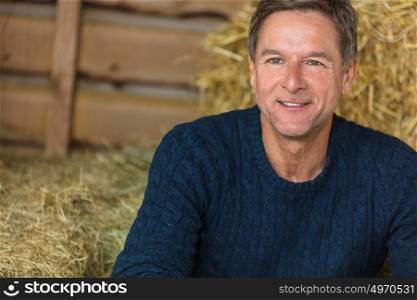 Portrait shot of an attractive, successful and happy middle aged man male wearing a blue sweater sitting on hay bales in a barn or stables
