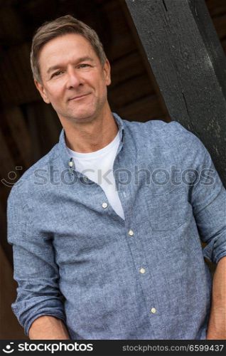 Portrait shot of an attractive, successful and happy middle aged man male wearing a blue shirt leaning on a post by a garage or barn