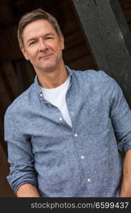 Portrait shot of an attractive, successful and happy middle aged man male wearing a blue shirt leaning on a post by a garage or barn