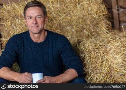 Portrait shot of an attractive, successful and happy middle aged man male wearing a blue sweater sitting on hay bales in a barn or stables drinking cup of tea or coffee