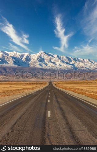 Portrait size shot of a straight empty highway leading to the snowy peaks of The Kuray mountain range. Beautiful blue cloudy sky as a background. Altai mountains, Siberia, Russia.. Portrait size shot of a straight empty highway leading to the snowy peaks of The Kuray mountain range. Beautiful blue cloudy sky as a background. Altai mountains, Siberia, Russia