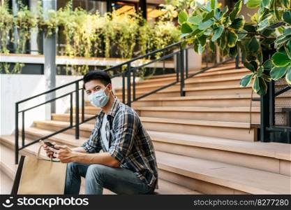 Portrait smiley Young handsome man in mask, holding paper bag and show credit card on stair of shopping mall, New normal lifestyle and shopping concept, copy space