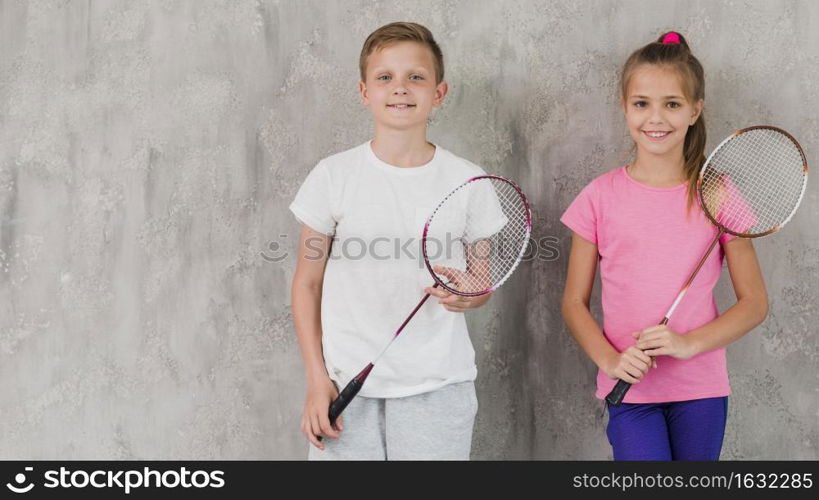 portrait smiling boy girl holding racket hands against concrete wall