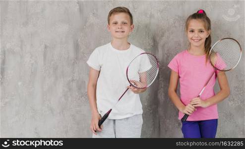 portrait smiling boy girl holding racket hands against concrete wall