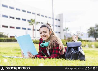 portrait smiling female university student lying green grass holding book hand