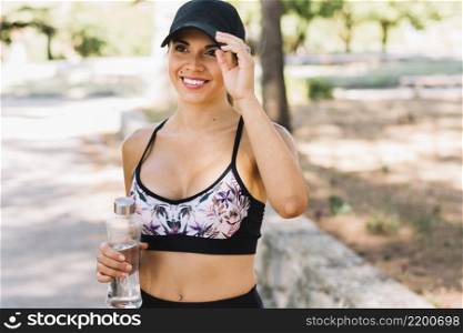 portrait smiling sporty young woman wearing black cap holding plastic water bottle