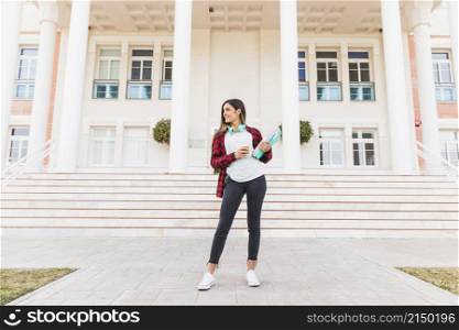 portrait smiling teenage female student holding books takeaway coffee cup standing front university building