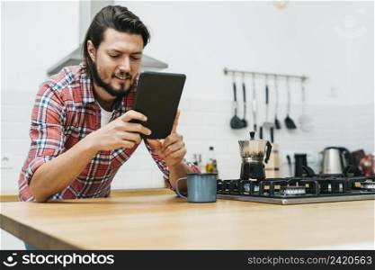 portrait smiling young man leaning wooden kitchen counter looking smart phone