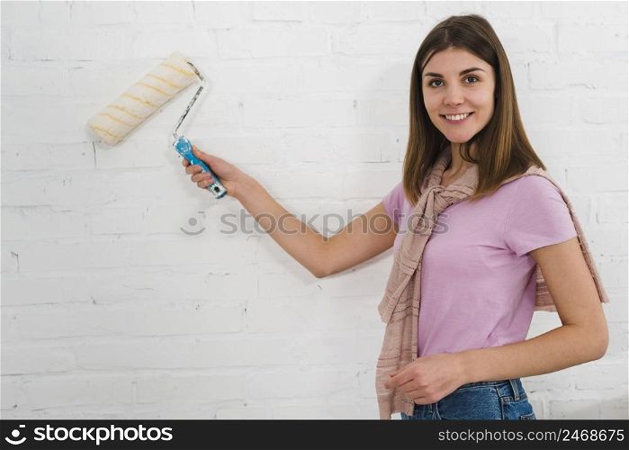portrait smiling young woman using paint roller white brick wall