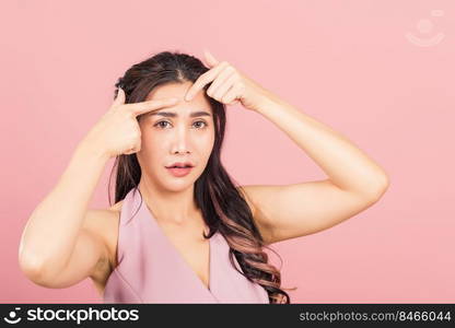 Portrait teenage Asian beautiful young woman having skin problems squeezing pimples on her face, studio shot on pink background, with copy space, Thai female acne, beauty care concept