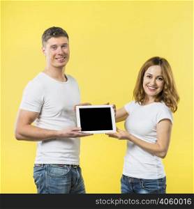 portrait two happy young couple showing black screen digital tablet against yellow backdrop