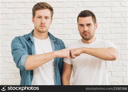 portrait two male friends bumping fist standing against white brick wall