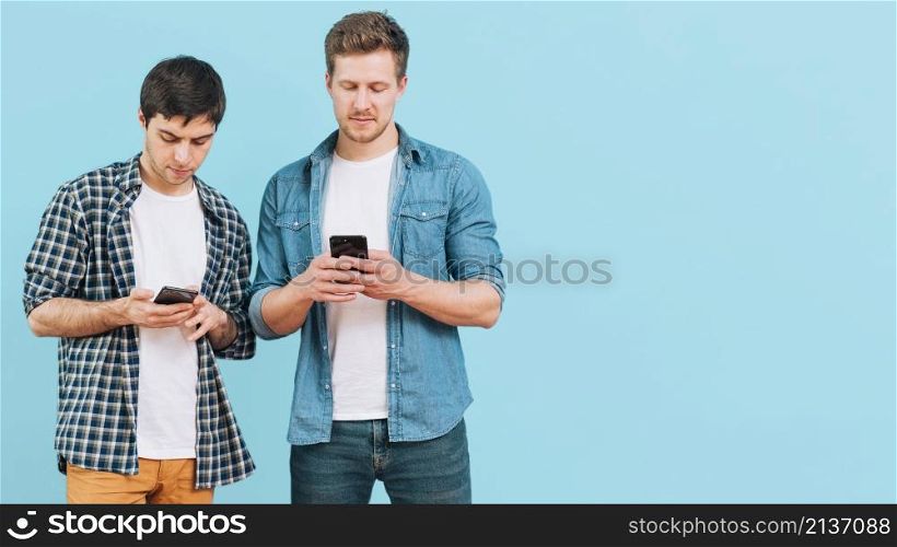 portrait two young men standing against blue background using mobile phone