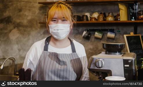 Portrait young Asia girl waitress wear medical face mask feeling happy smile waiting for clients after lockdown at urban cafe. Owner small business, food and drink, business reopen again concept.