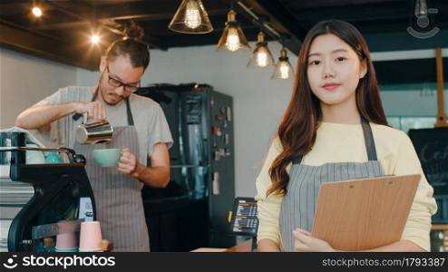 Portrait young Asian couple barista feeling happy with menu at urban cafe. Small business owner Korean friends in apron relax toothy smile looking to camera standing at the counter in coffee shop.