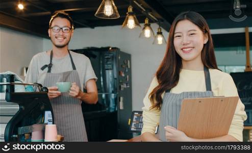 Portrait young Asian couple barista feeling happy with menu at urban cafe. Small business owner Korean friends in apron relax toothy smile looking to camera standing at the counter in coffee shop.