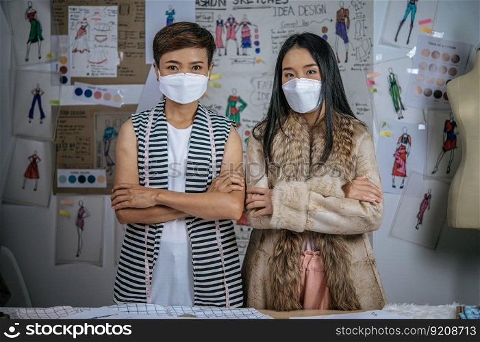 Portrait Young Asian female and friend standing with cross arms together in modern atelier. Positive women creative coworkers wearing medical face mask in studio. distance and new normal concept
