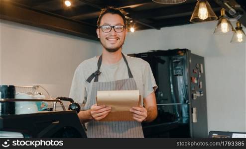 Portrait young Asian guy barista take receive order with menu feeling happy at urban cafe. Asia small business owner boy in apron relax toothy smile looking to camera stand at counter in coffee shop.