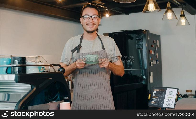 Portrait young Asian guy barista waitress holding coffee cup feeling happy at urban cafe. Asia small business owner boy in apron relax toothy smile looking to camera stand at counter in coffee shop.