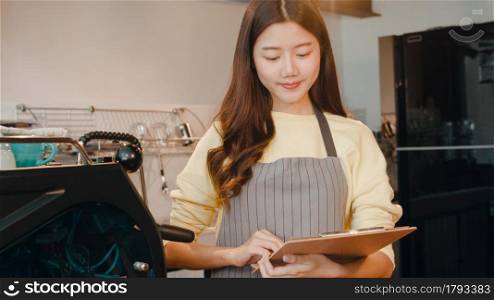 Portrait young Asian lady barista take receive order with menu feeling happy at urban cafe. Asia small business owner girl in apron relax toothy smile looking to camera stand at counter in coffee shop