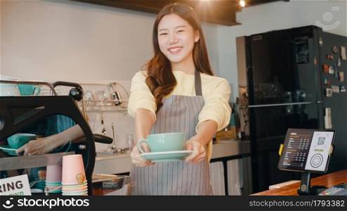 Portrait young Asian lady barista waitress holding coffee cup feeling happy at urban cafe. Asia small business owner girl in apron relax toothy smile looking to camera stand at counter in coffee shop.