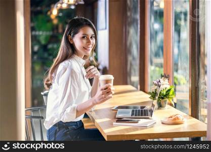 Portrait Young Asian woman holding and drinking a cup of coffee and working with technology laptop at a coffee shop. freelancer and entrepreneur working by connecting to internet via computer
