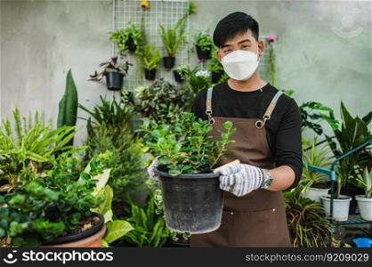 Portrait Young gardener man in face mask and wearing gloves and apron, stand and hold houseplants in pot and looking to camera, small business with tree or Home gardening
