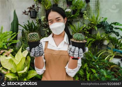 Portrait Young gardener woman in face mask wearing gloves and apron show two cactus in hand, houseplants in pot behind him, small business with tree or  Home gardening
