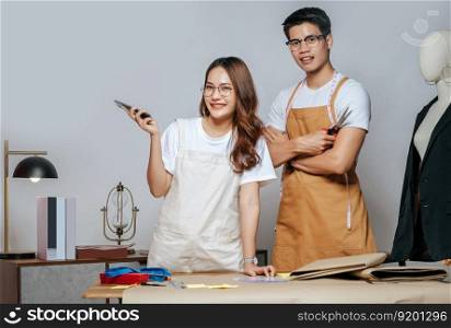 Portrait Young handsome man and pretty woman, Couple fashion designer working with smartphone, paper clothing pattern on desk at the studio at home