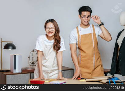 Portrait Young handsome man and pretty woman in glasses wearing apron, Couple fashion designer working together with happy, mannequin and tailoring tools on desk at studio