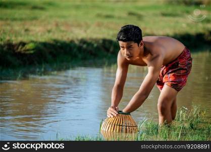 Portrait Young man topless use bamboo fishing trap to catch fish for cooking, Asian young farmer man in rural lifestyle, blurred buffalo is soaking in the sw&. in background