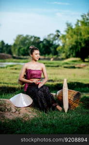 Portrait Young pretty Asian woman in beautiful Thai traditional clothes at rice field, she sitting near fishing equipment, bamboo trap, trap shrimp, and spoon-net