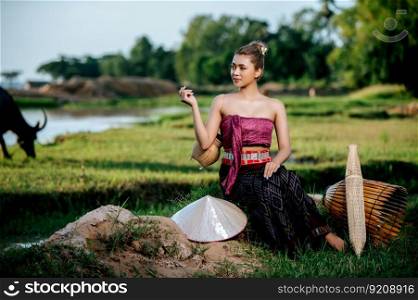 Portrait Young pretty Asian woman in beautiful Thai traditional clothes at rice field, she sitting near fishing equipment, bamboo trap, trap shrimp, and spoon-net
