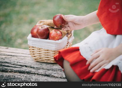 Portrait young woman with Little Red Riding Hood costume with apple and bread on basket sitting in green tree park background