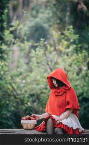 Portrait young woman with Little Red Riding Hood costume with apple and bread on basket sitting in green tree park background