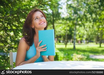 Positive adult beautiful woman sitting near table and reading book outdoors at backyard in summer