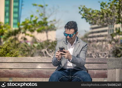 Positive adult male entrepreneur in classy outfit and sunglasses sitting on bench on urban street and smiling happily while getting great news on smartphone. Happy businessman reading message on smartphone on street
