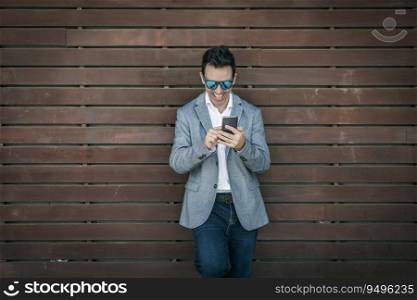 Positive adult man in trendy outfit and sunglasses leaning back on wooden wall and messaging on mobile phone. Happy stylish businessman messaging on smartphone