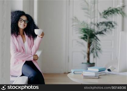 Positive African American female business worker sits at windowsill, holds mug of coffee, dressed in formal clothes, poses near desktop with books, green indoor plant in background, has break