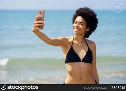 Positive African American female in swimwear taking self portrait on smartphone while standing on shore near waving sea on sunny day in summer. Black woman in bikini taking selfie near sea