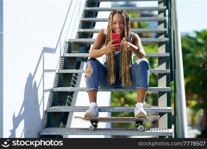 Positive African American female skater with braids and skateboard sitting on stairs and messaging on social media on cellphone in summer. Cheerful black female skater browsing smartphone in city