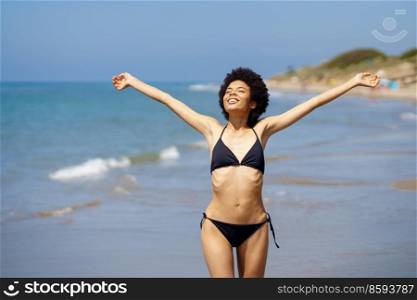 Positive African American female tourist in swimwear walking on coast with outstretched arms near sea on summer day in tropical resort and enjoying freedom. Delighted black woman in bikini walking on beach