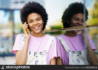 Positive African American female with Afro hairstyle looking away while having phone conversation near glass building in city. Cheerful black woman talking on smartphone near building