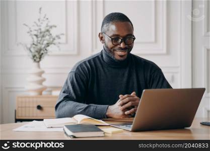 Positive African American male private consultant having session online listens to client attentively while looking at computer screen with smile, working remotely from home office. Freelance concept. Positive African American male private consultant having session online listens to client attentively