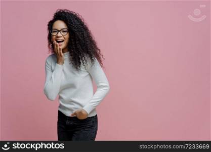 Positive African American woman laughs at something funny, covers opened mouth with palm, cannot control her emotions, keeps hand in pocket of jeans, wears white poloneck, isolated on pink background