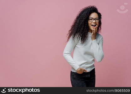 Positive African American woman laughs at something funny, covers opened mouth with palm, cannot control her emotions, keeps hand in pocket of jeans, wears white poloneck, isolated on pink background