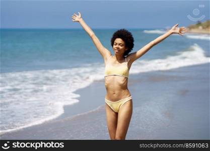 Positive African American woman opening her arms on the beach to enjoy her holiday in the sun. Young black female wearing yellow bikini.. Positive black woman opening her arms on the beach to enjoy her holiday in the sun.