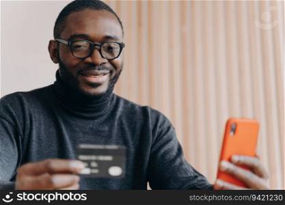 Positive African entrepreneur in glasses, paying online with credit card, ordering on cellphone, smiling millennial guy, using mobile banking app for transaction.